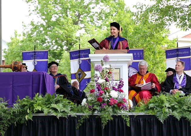 Dr. Nichole DeWall, associate professor of English at McKendree University, addresses the crowd at the 2018 McKendree commencement ceremony as winner of the Grandy Faculty Award. Looking on, left to right, is Dr. Christine Bahr, provost and Dean; Dr. James Dennis, university president;  and Daniel Lett, president of the board of trustees.